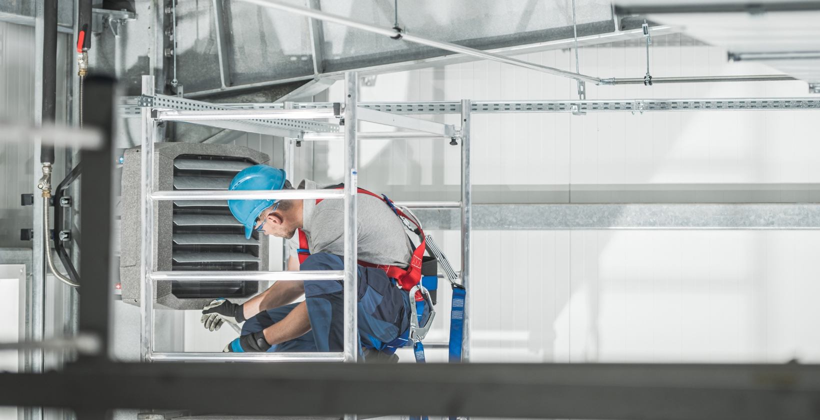 Emergency gas repairs UK, 24/7 Engineer in a hard hat and safety harness repairing a wall-mounted warehouse gas heater from an access tower in the UK
